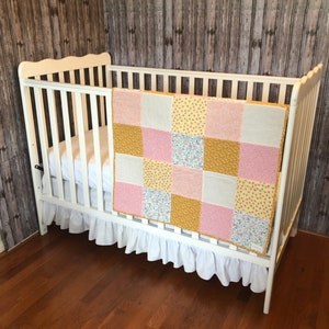 Puede incluir: Una cuna blanca con una falda de cuna blanca con volantes y una colcha de retazos en tonos rosa, amarillo y blanco. La colcha tiene un patrón floral.