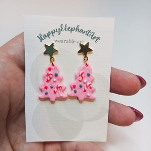 May include: Pink Christmas tree earrings with gold star tops and candy cane and sprinkle details. The earrings are on a white card with the text "HappyElephantArt wearable art".