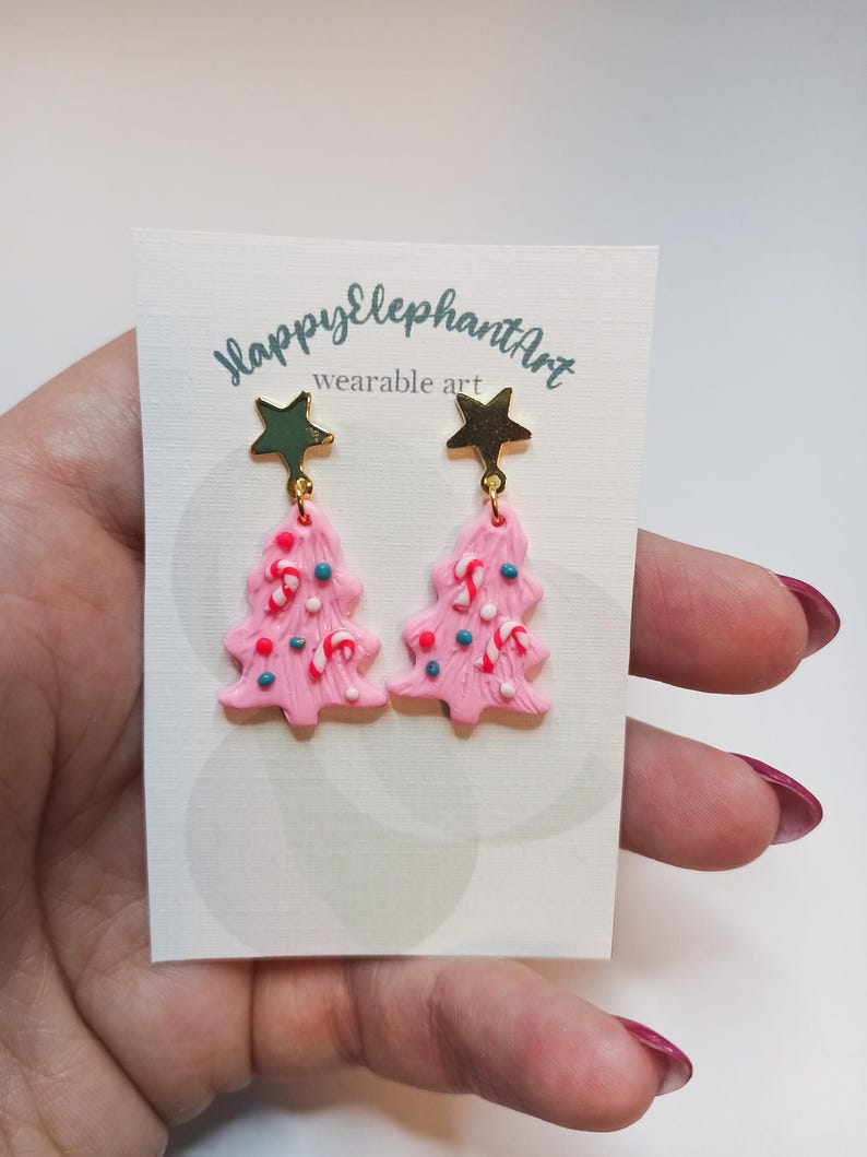May include: Pink Christmas tree earrings with gold star tops and candy cane and sprinkle details. The earrings are on a white card with the text "HappyElephantArt wearable art".