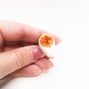 May include: A miniature, realistic-looking egg, cut in half, with a white egg white and a bright orange yolk. The yolk is speckled with red and brown. The egg is held against a white background.