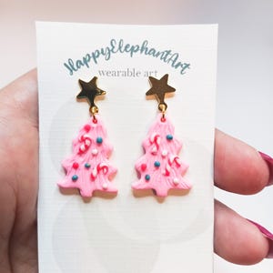 May include: Pink Christmas tree earrings with gold star charms. The trees are decorated with white, red, and blue sprinkles and candy canes. The earrings are labeled "Happy Elephant Art wearable art".