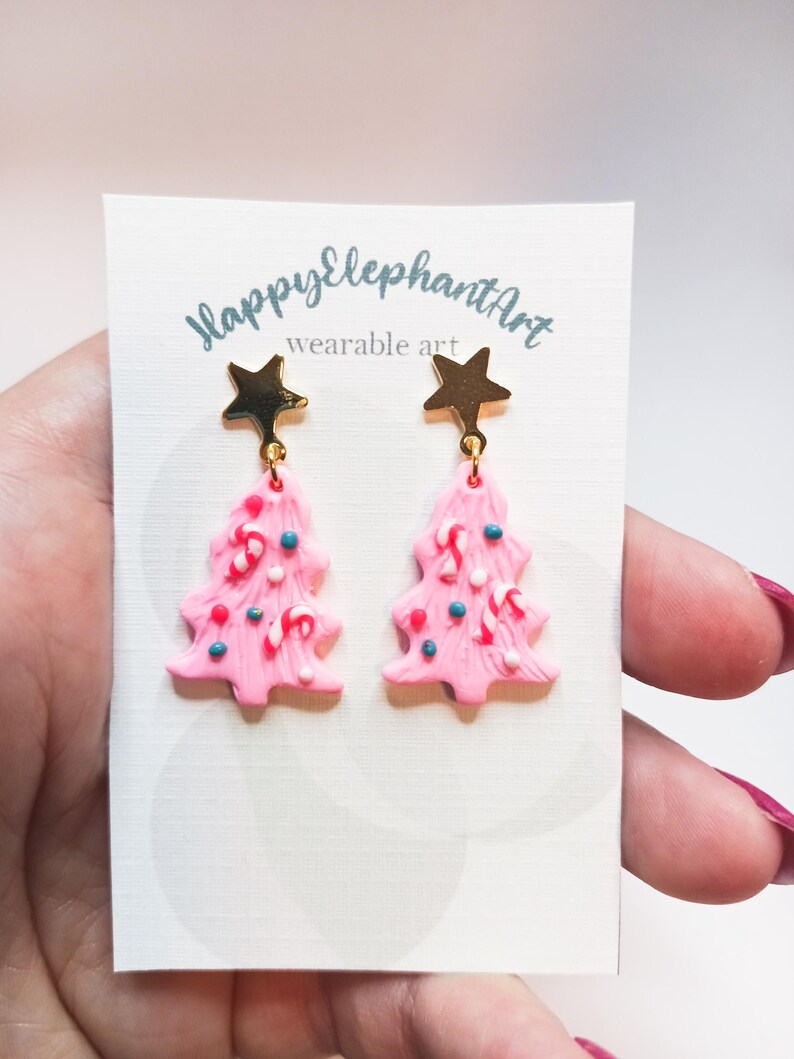May include: Pink Christmas tree earrings with gold star charms. The trees are decorated with white, red, and blue sprinkles and candy canes. The earrings are labeled "Happy Elephant Art wearable art".