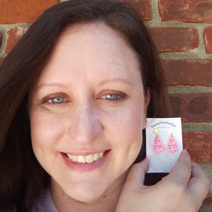May include: A pair of pink Christmas tree earrings with gold star accents. The earrings are on a white card with the text "Happy Elephant" and "Handmade with Love".