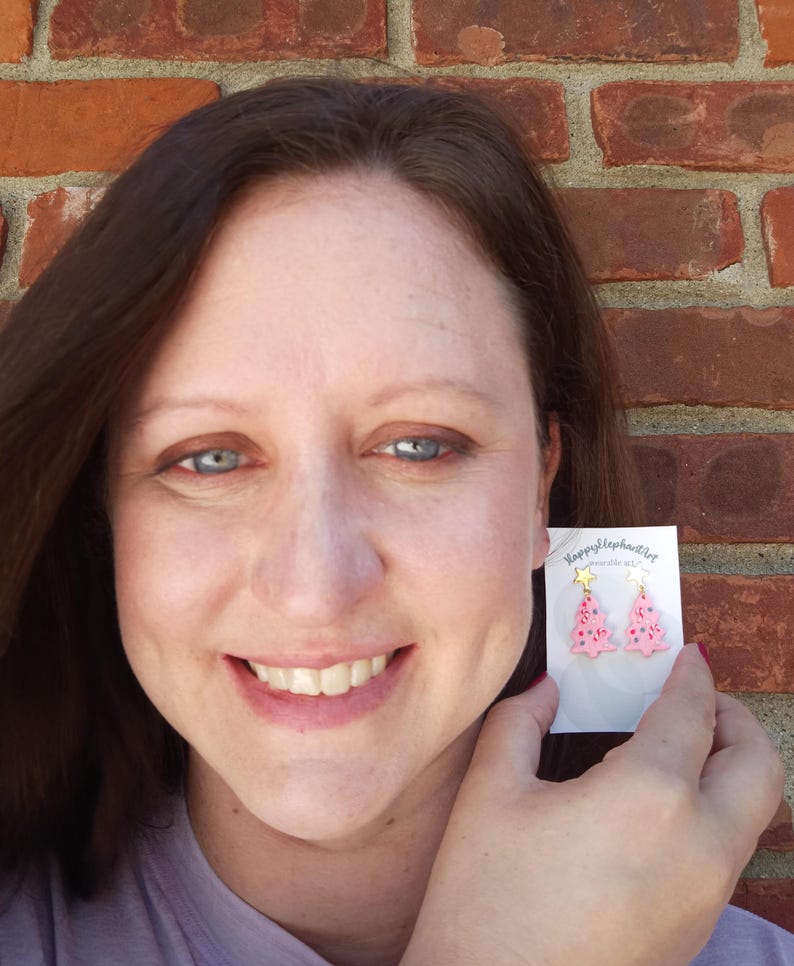 May include: A pair of pink Christmas tree earrings with gold star accents. The earrings are on a white card with the text "Happy Elephant" and "Handmade with Love".
