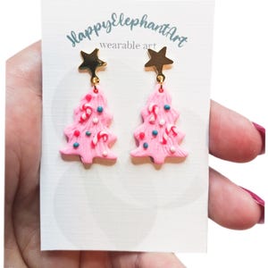 May include: A pair of pink Christmas tree earrings with gold star charms. The trees are decorated with white sprinkles, red candy canes, and blue and white swirls. The earrings are on a white card with the text "Happy Elephant Art wearable art".