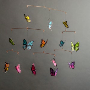 May include: A copper-toned mobile featuring colorful butterfly cutouts. The butterflies are in shades of yellow, green, blue, orange, pink, and purple. The mobile is suspended by thin wires, creating a delicate and airy display.