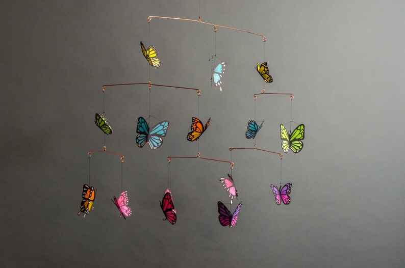May include: A copper-toned mobile featuring colorful butterfly cutouts. The butterflies are in shades of yellow, green, blue, orange, pink, and purple. The mobile is suspended by thin wires, creating a delicate and airy display.