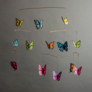 May include: A mobile featuring colorful butterfly decorations. The butterflies are in shades of yellow, blue, green, orange, pink, and purple. The butterflies are suspended from copper-colored metal rods.