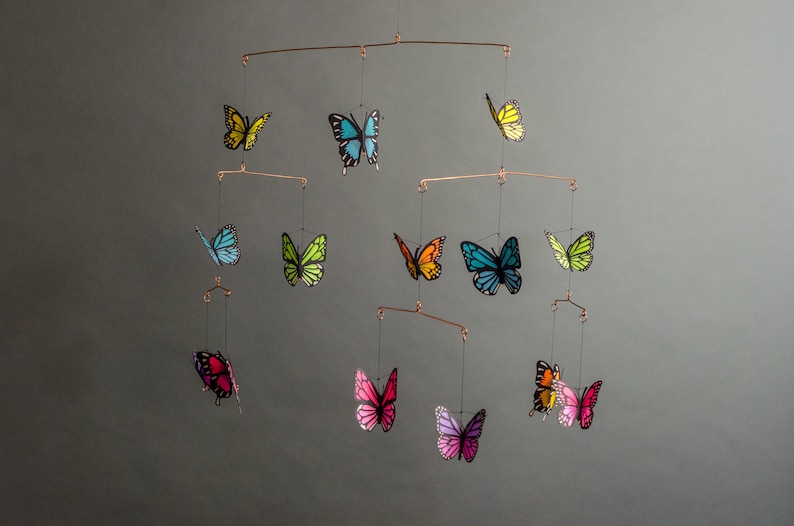May include: A mobile featuring colorful butterfly decorations. The butterflies are in shades of yellow, blue, green, orange, pink, and purple. The butterflies are suspended from copper-colored metal rods.