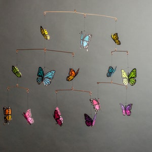 May include: A handcrafted mobile featuring colorful butterfly cutouts in various sizes. The butterflies are in shades of yellow, blue, green, orange, pink, and purple, suspended from copper-colored wire. A decorative accent for a nursery or living space.