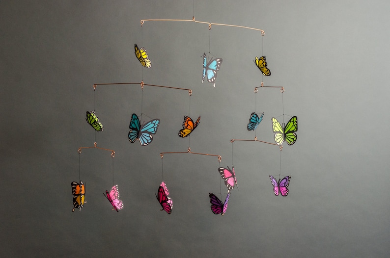 May include: A handcrafted mobile featuring colorful butterfly cutouts in various sizes. The butterflies are in shades of yellow, blue, green, orange, pink, and purple, suspended from copper-colored wire. A decorative accent for a nursery or living space.