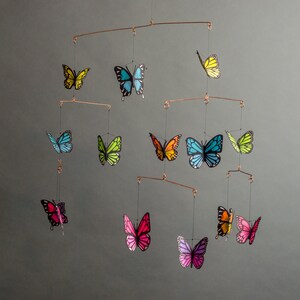 May include: A mobile featuring colorful butterfly cutouts in shades of yellow, blue, green, orange, pink, and purple. The butterflies are suspended from copper-colored metal rods, creating a decorative hanging art piece. The background is a solid gray.