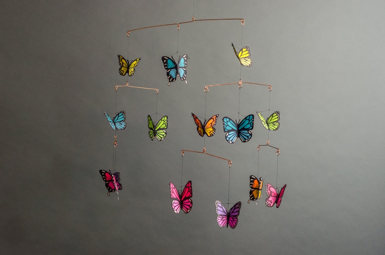 May include: A mobile featuring colorful butterfly cutouts in shades of yellow, blue, green, orange, pink, and purple. The butterflies are suspended from copper-colored metal rods, creating a decorative hanging art piece. The background is a solid gray.
