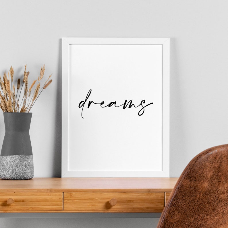 May include: A white framed print with the word "dreams" written in black cursive script. The print is hanging on a white wall above a wooden desk with two drawers.