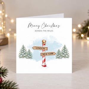 May include: A square white greeting card featuring the text Merry Christmas ACROSS THE MILES. The design includes a red and white striped pole with two wooden signs pointing to London and New York, surrounded by four green pine trees in snow.
