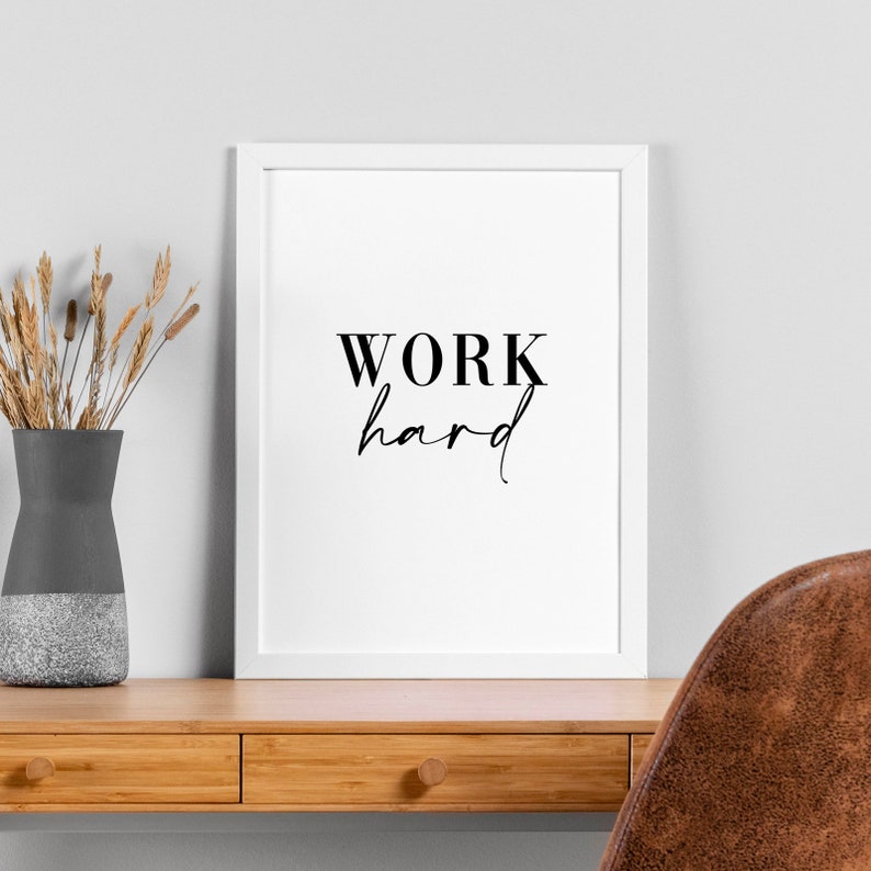 Op de afbeelding: Een wit ingelijst print met de tekst "WORK hard" in zwarte letters. De print hangt aan een witte muur boven een houten bureau met een bruine leren stoel.
