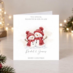 May include: A square white greeting card featuring an illustration of two snowmen wearing red knit hats and scarves. The text reads TO A SPECIAL DAUGHTER & SON IN LAW, Rachel & James, and MERRY CHRISTMAS. The snowmen have orange carrot noses.