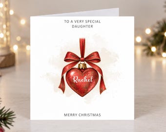 May include: A white square greeting card featuring the text TO A VERY SPECIAL DAUGHTER at the top and MERRY CHRISTMAS at the bottom. In the center is a red heart-shaped Christmas ornament with a red ribbon bow and the name Rachel.