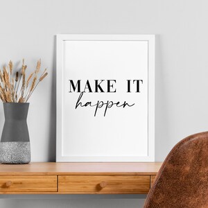Op de afbeelding: Een wit ingelijst print met de tekst "MAKE IT happen" in zwarte letters. De print staat op een houten bureau met een vaas met gedroogde bloemen en een bruine leren stoel.
