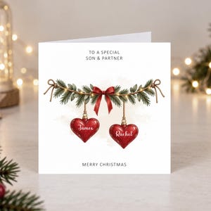 May include: A white square greeting card featuring the text TO A SPECIAL SON & PARTNER at the top and MERRY CHRISTMAS at the bottom. The center shows a pine branch garland with a red bow and two red heart ornaments labeled James and Rachel.