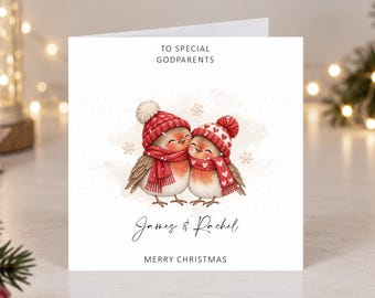 Könnte beinhalten: Eine quadratische weiße Grußkarte mit einer Illustration von zwei Rotkehlchen, die rote Strickmützen und Schals tragen. Der Text lautet TO SPECIAL GODPARENTS, James & Rachel und MERRY CHRISTMAS. Die Vögel sind von kleinen Schneeflocken umgeben.