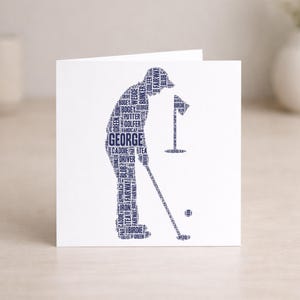 May include: A white greeting card featuring a navy blue silhouette of a golfer in action. The golfer's form is composed of golf-related words, with "GEORGE" prominently displayed. A golf ball and flag are also visible.
