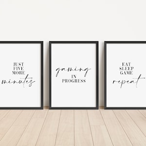 May include: Three black framed prints with white backgrounds and black text. The left print says "Just five more minutes". The middle print says "Gaming in progress". The right print says "Eat sleep game repeat".