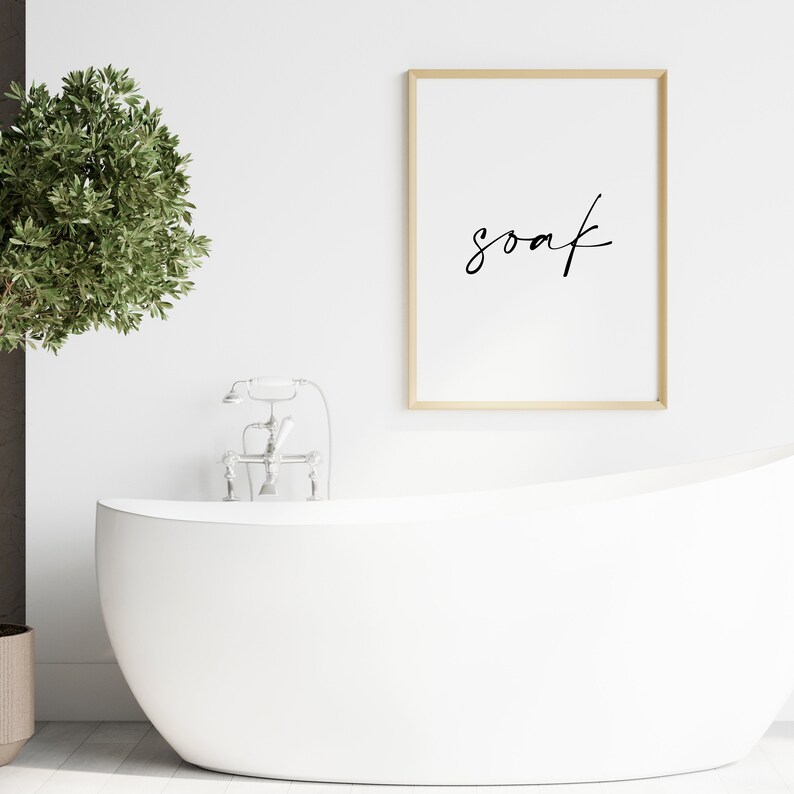 May include: A white bathtub with a silver faucet and a framed print with the word "soak" in black calligraphy. The print is hanging on a white wall.