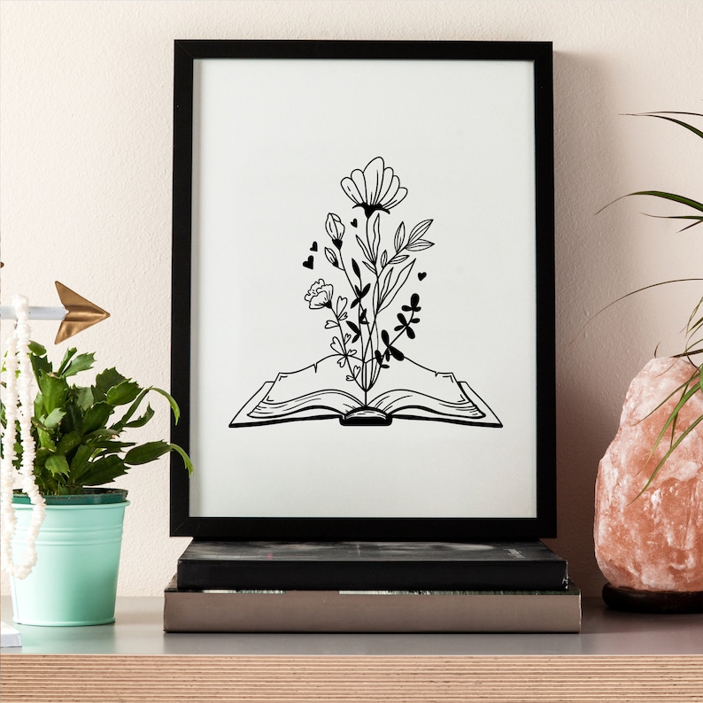May include: Black and white line art print of an open book with flowers growing out of it. The book is in the center of the image and the flowers are growing upwards. The print is framed in a black frame.