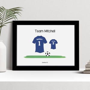 K&ouml;nnte beinhalten: Gerahmter Druck mit Fu&szlig;ballmotiv. Das Kunstwerk zeigt zwei blaue Fu&szlig;balltrikots, eines mit der Aufschrift "Daddy 1" und das andere mit "Jacob 2", einen Fu&szlig;ball und den Text "Team Mitchell". Der Text unten lautet "Chelsea FC".