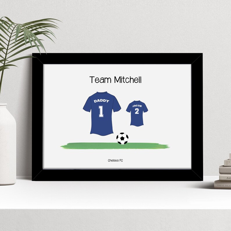 K&ouml;nnte beinhalten: Gerahmter Druck mit Fu&szlig;ballmotiv. Das Kunstwerk zeigt zwei blaue Fu&szlig;balltrikots, eines mit der Aufschrift "Daddy 1" und das andere mit "Jacob 2", einen Fu&szlig;ball und den Text "Team Mitchell". Der Text unten lautet "Chelsea FC".