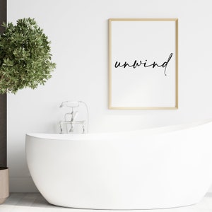 May include: A white bathtub with a silver faucet and a framed print with the word "unwind" in black script on a white background.