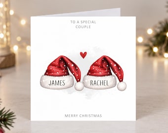 Könnte beinhalten: Eine quadratische weiße Grußkarte mit zwei roten Weihnachtsmützen mit weißem Fellbesatz und Bommeln. Die Mützen sind mit den Namen James und Rachel personalisiert. Über den Mützen befindet sich ein kleines rotes Herz sowie der Text To A Special Couple und Merry Christmas.