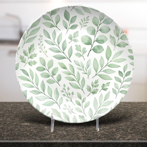 Può includere: Un piatto in ceramica bianca con un motivo a foglie verdi. Il piatto è rotondo e presenta vari disegni di foglie in tonalità di verde. Il piatto è esposto su un supporto trasparente, mostrando un design ispirato alla natura.