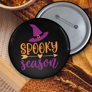 May include: A black button with the words "Spooky Season" in orange and purple, featuring a witch hat and arrow design. The button is set against a backdrop of autumn leaves and a cozy, textured surface.