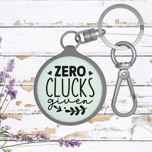 May include: A silver key chain with a round, light green charm that says "ZERO CLUCKS given" in black lettering. The charm has a silver border and two small hearts on either side of the text.