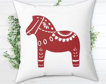 May include: White square pillow featuring a red Dala horse design. The horse is decorated with white floral and geometric patterns. The pillow is a decorative item, suitable for home decor.