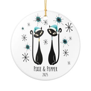 Op de afbeelding: Een ronde, witte kerstversiering met twee zwarte katten met blauwe mutsen. De versiering heeft een gouden ophangkoord en de tekst "PIXIE & PEPPER 2025" eronder. Sneeuwvlokken sieren het ontwerp.