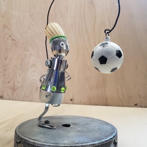 May include: A silver robot with a wooden head and green eyes is holding a black and white football with a string. The robot is standing on a black circular base with five star cutouts.