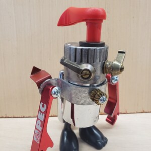 May include: A handcrafted robot sculpture made from recycled metal components. It has a red handle, red bottle opener arms with "PEC" text, a silver body, and black feet. The robot stands on a light wood surface.