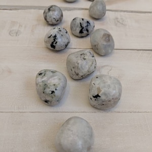 May include: A collection of ten white and gray tumbled moonstone stones with black flecks. The stones are arranged in a line on a light wood surface.