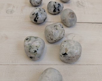May include: A collection of ten white and gray tumbled moonstone stones with black flecks. The stones are arranged in a line on a light wood surface.