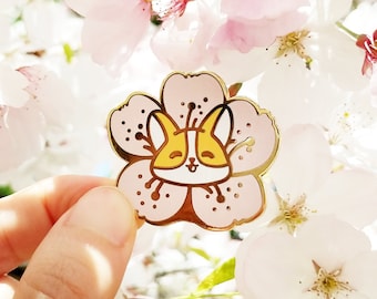 Peut inclure: Une épingle en émail dorée représentant un renard de dessin animé à l'intérieur d'une fleur de cerisier rose. L'épingle est tenue dans une main sur un fond de fleurs de cerisier blanches et roses.