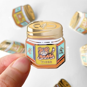 May include: Gold-colored enamel pin shaped like a jar of Tiger Balm with a tiger illustration and Chinese characters. The jar is yellow, orange, blue, and white.