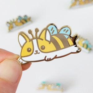 May include: Gold-tone enamel pin featuring a cartoon bee with a yellow and black striped body, blue wings, and a white face. The bee is lying on its side with its legs in the air.