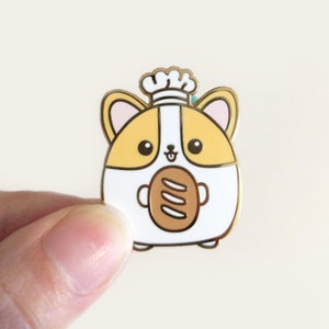 May include: A cute enamel pin featuring a corgi chef holding a loaf of bread. The pin has a gold outline, with the corgi's body in white and yellow. The chef's hat is white with a gold trim. The bread is brown.