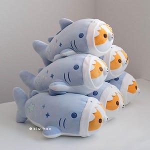 May include: A stack of plush toys shaped like blue sharks with a white belly and a brown and orange corgi head poking out of the shark's mouth. The plush toys have embroidered details, including stars and plus signs.