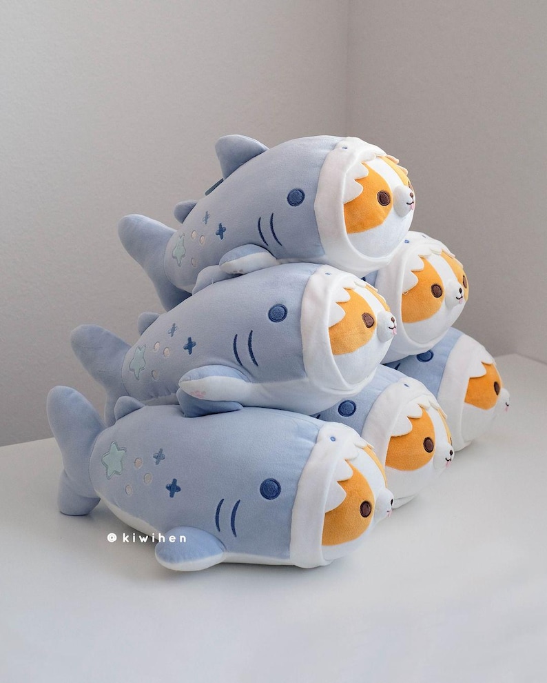 May include: A stack of plush toys shaped like blue sharks with a white belly and a brown and orange corgi head poking out of the shark's mouth. The plush toys have embroidered details, including stars and plus signs.