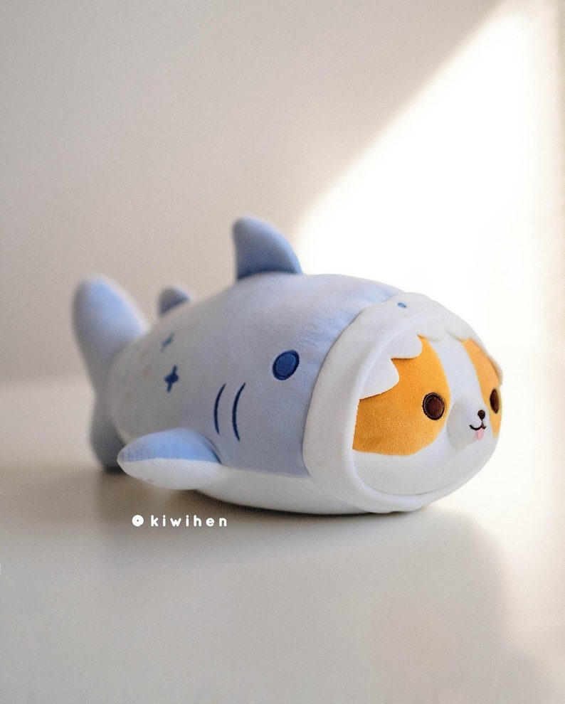May include: A plush toy shaped like a shark with a cute dog face peeking out from the shark's mouth. The shark is light blue with white accents and the dog is orange and white.