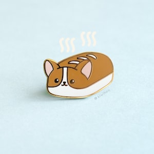 May include: A gold-colored enamel pin depicting a brown and white cartoon dog shaped like a loaf of bread with steam coming off of it. The pin says "kiwiihen" at the bottom.